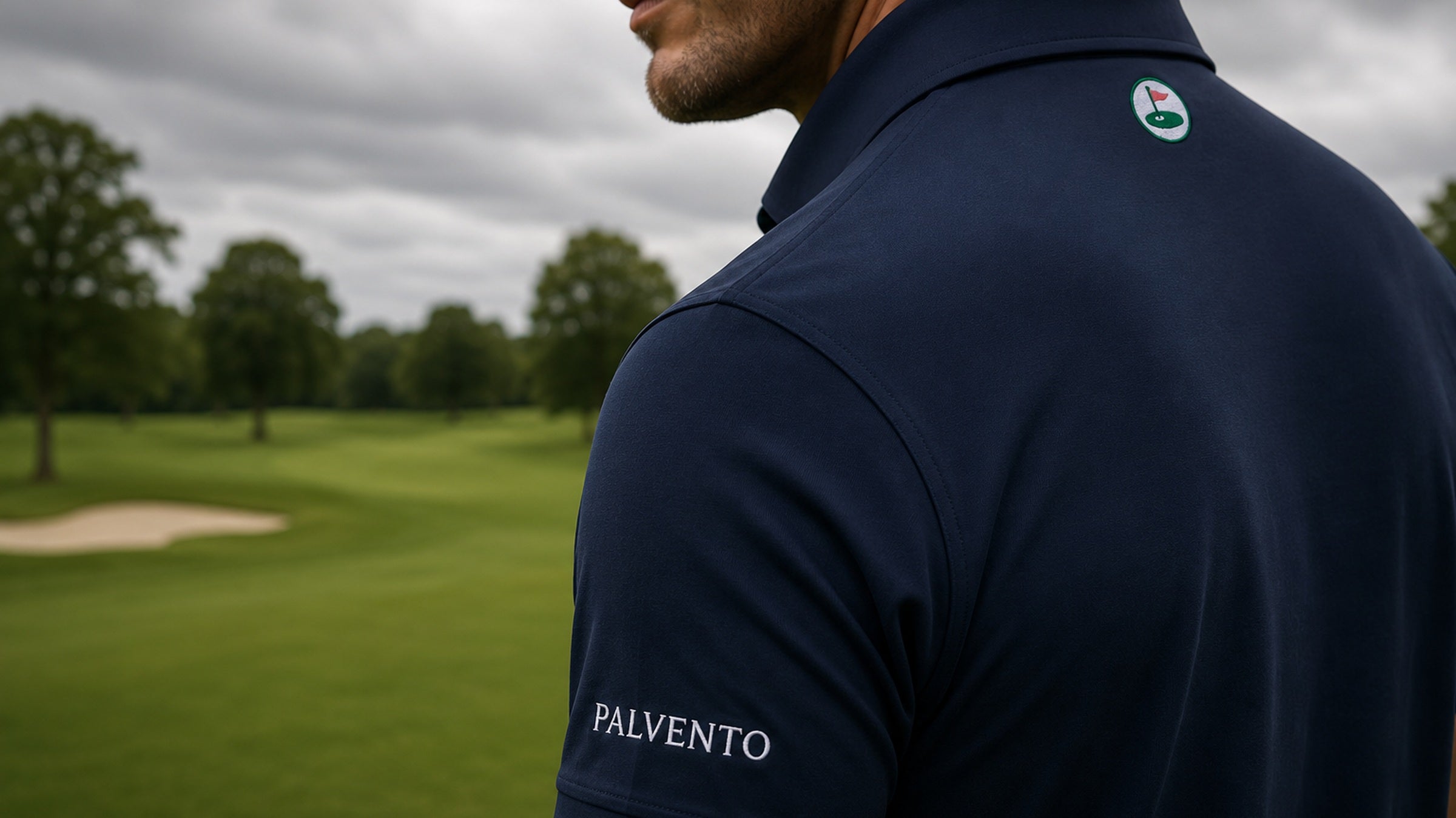 Man wearing a navy Palvento Golf polo on a parkland golf course, showing the rear roundel and sleeve wordmark.
